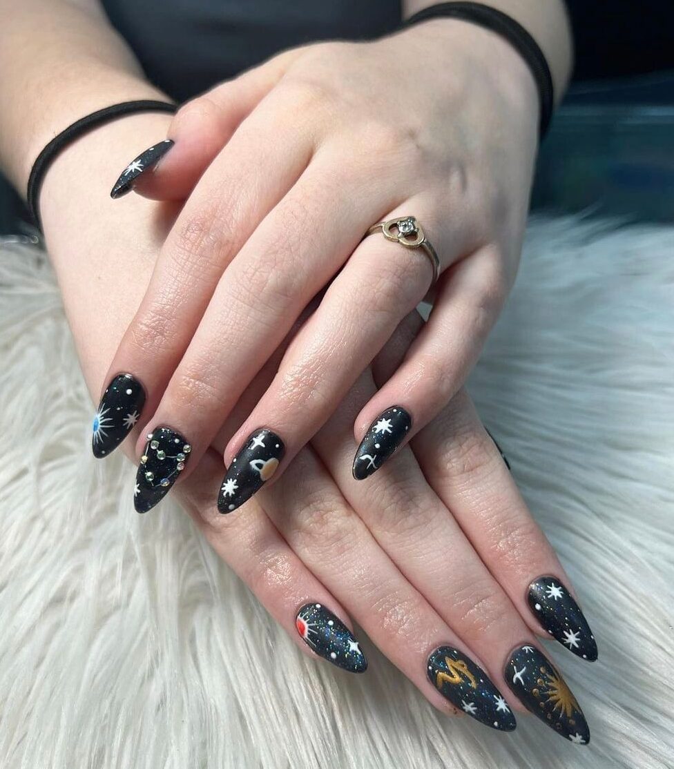 Vernis à ongles noir scintillant avec symbole du Capricorne et motifs d’ongles d’éléments célestes sur de longs ongles en amande