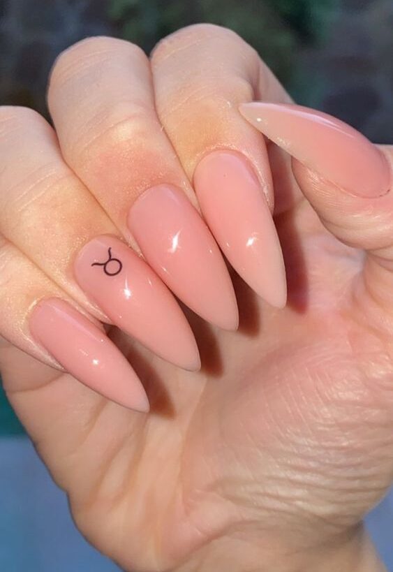 Couleur d’ongle nude avec un nail art minimaliste du signe Taureau sur de longs ongles en acrylique en forme d’amande