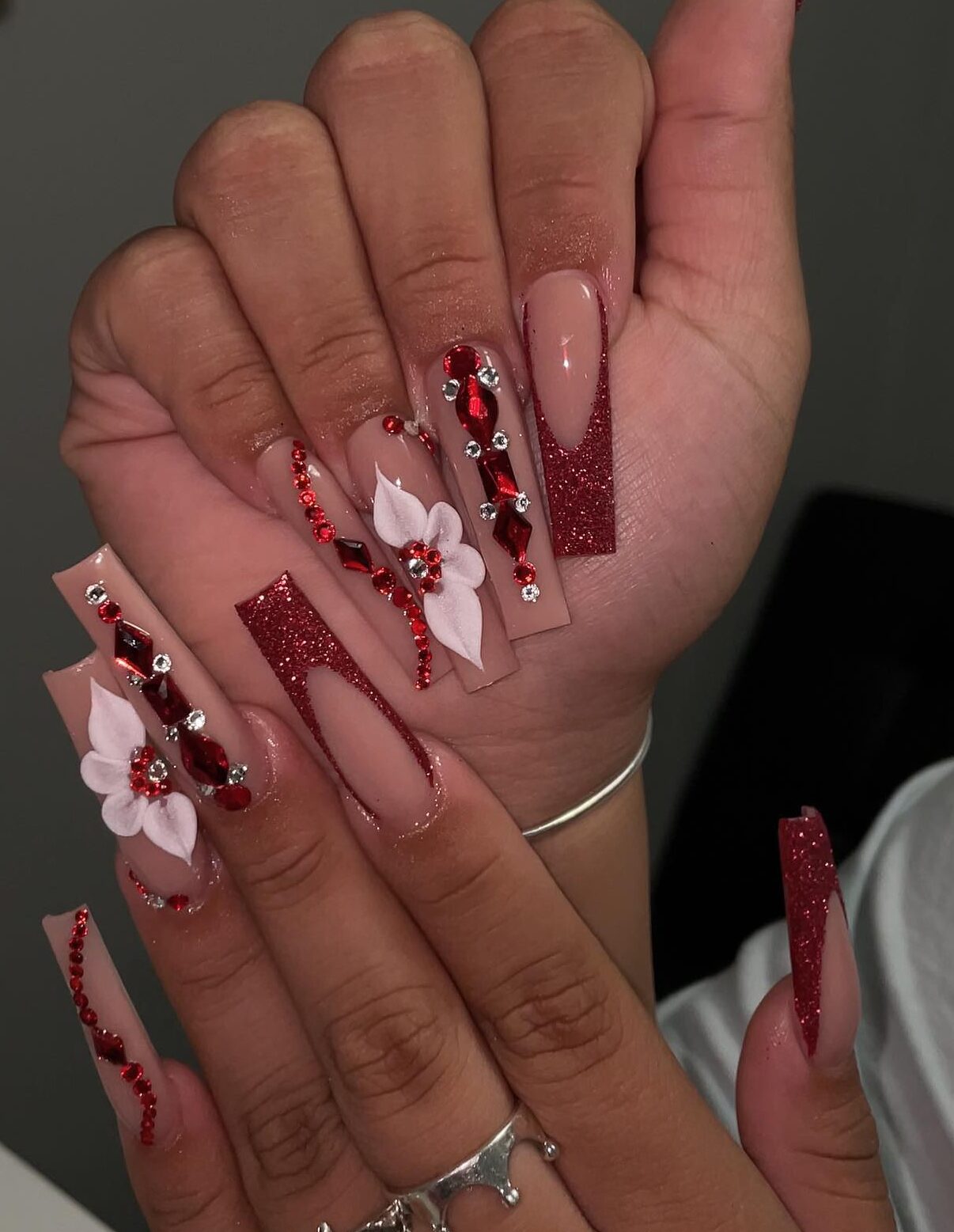 Vernis à ongles nude avec des pointes françaises scintillantes rouges, des strass et des fleurs 3D sur de longs ongles carrés