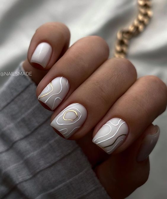 Vernis à ongles de couleur blanche au fini mat avec des motifs tourbillonnants blancs et dorés sur des ongles carrés courts et effilés